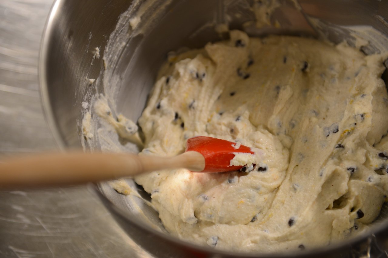 A red spatula stirs thick cookie dough with chocolate chips in a metal mixing bowl.