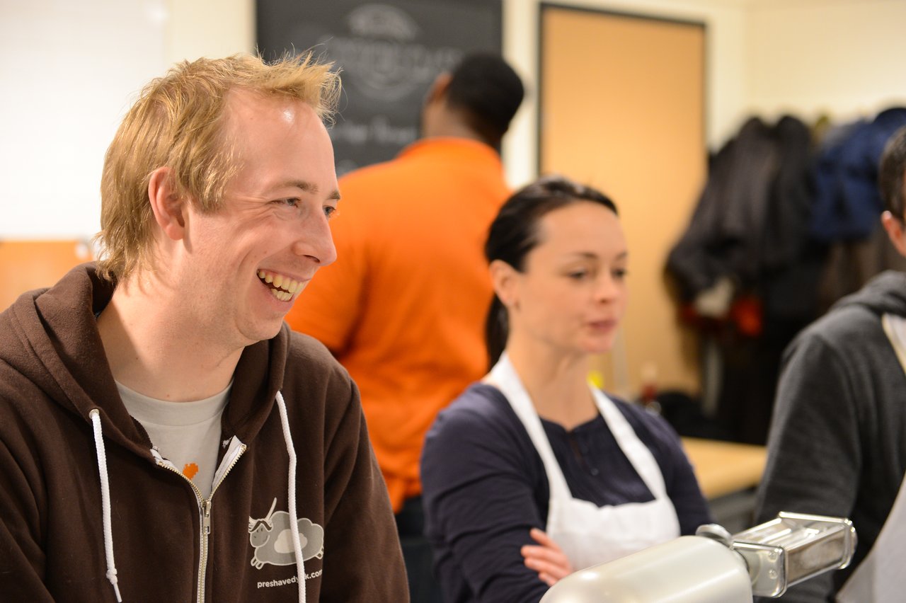 People participating in a cooking class, with one person smiling and another focused near a pasta-making machine.