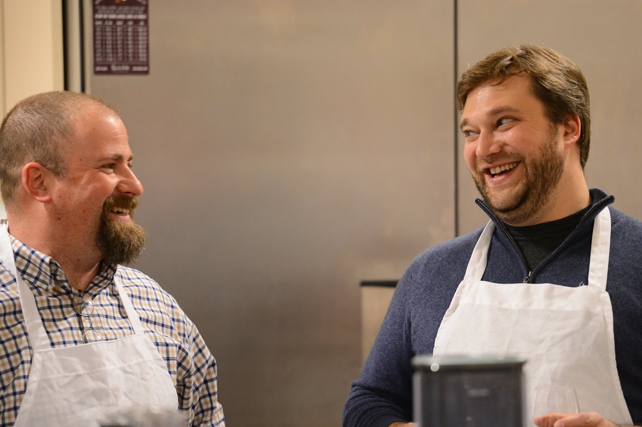 Two men wearing aprons smile and laugh while talking in a kitchen during a cooking class.