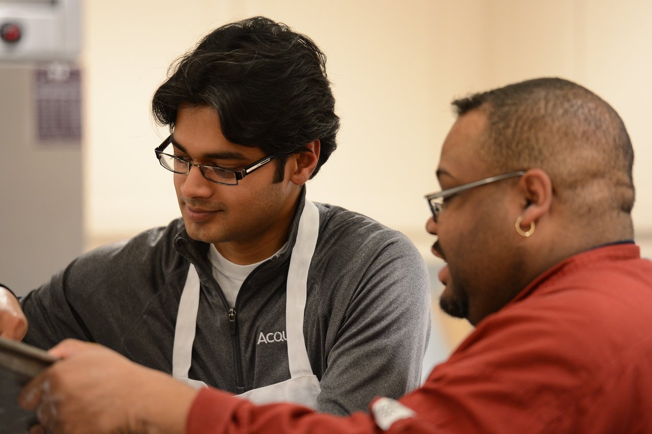 Two people in a cooking class, one wearing an apron and listening while the other gives instructions.