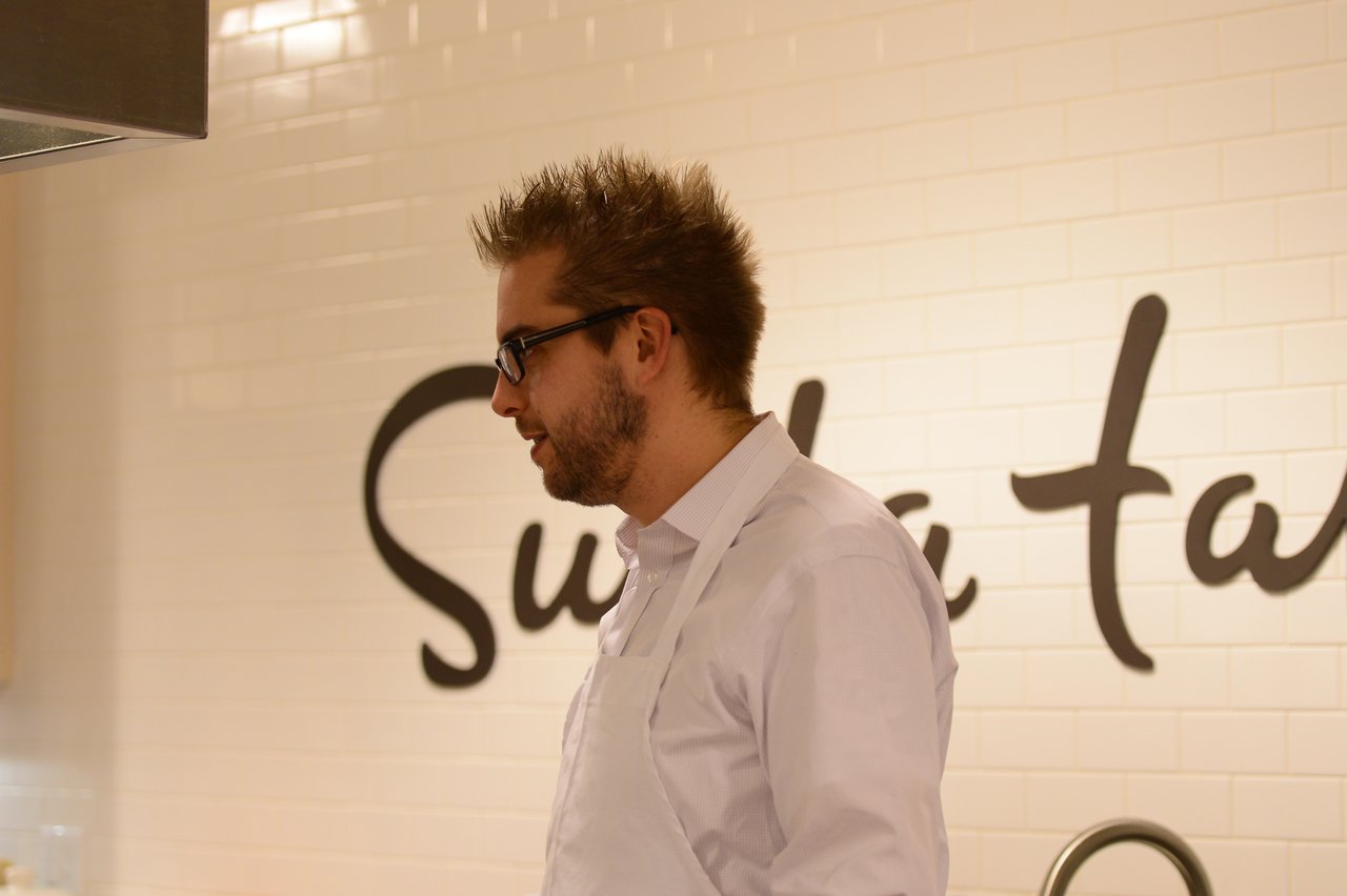 A man wearing glasses and a white apron stands in a kitchen, possibly participating in a cooking class.