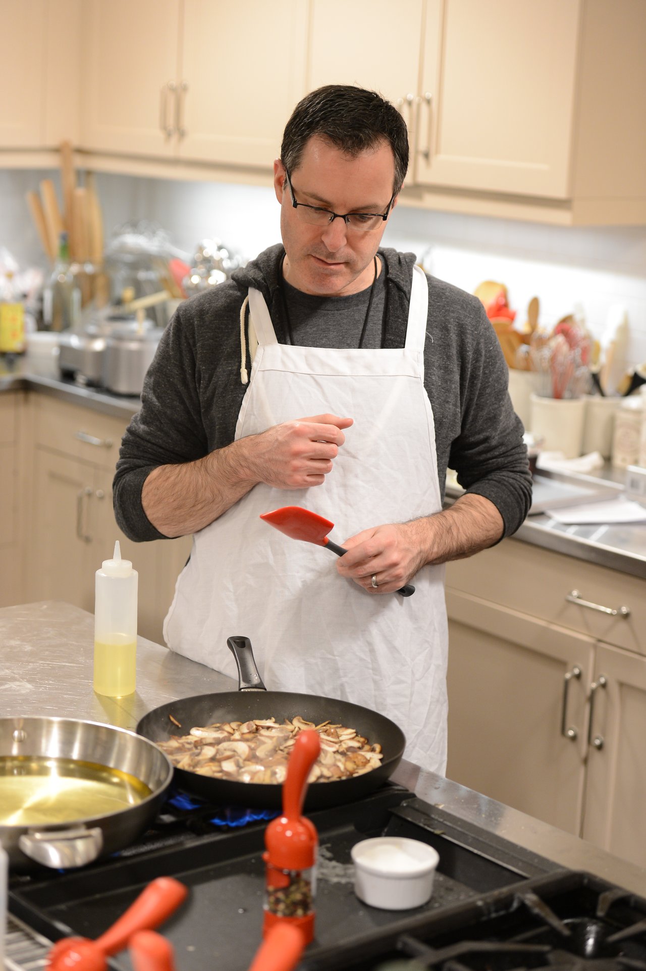 A person wearing an apron holds a spatula while cooking mushrooms in a pan on a stovetop.