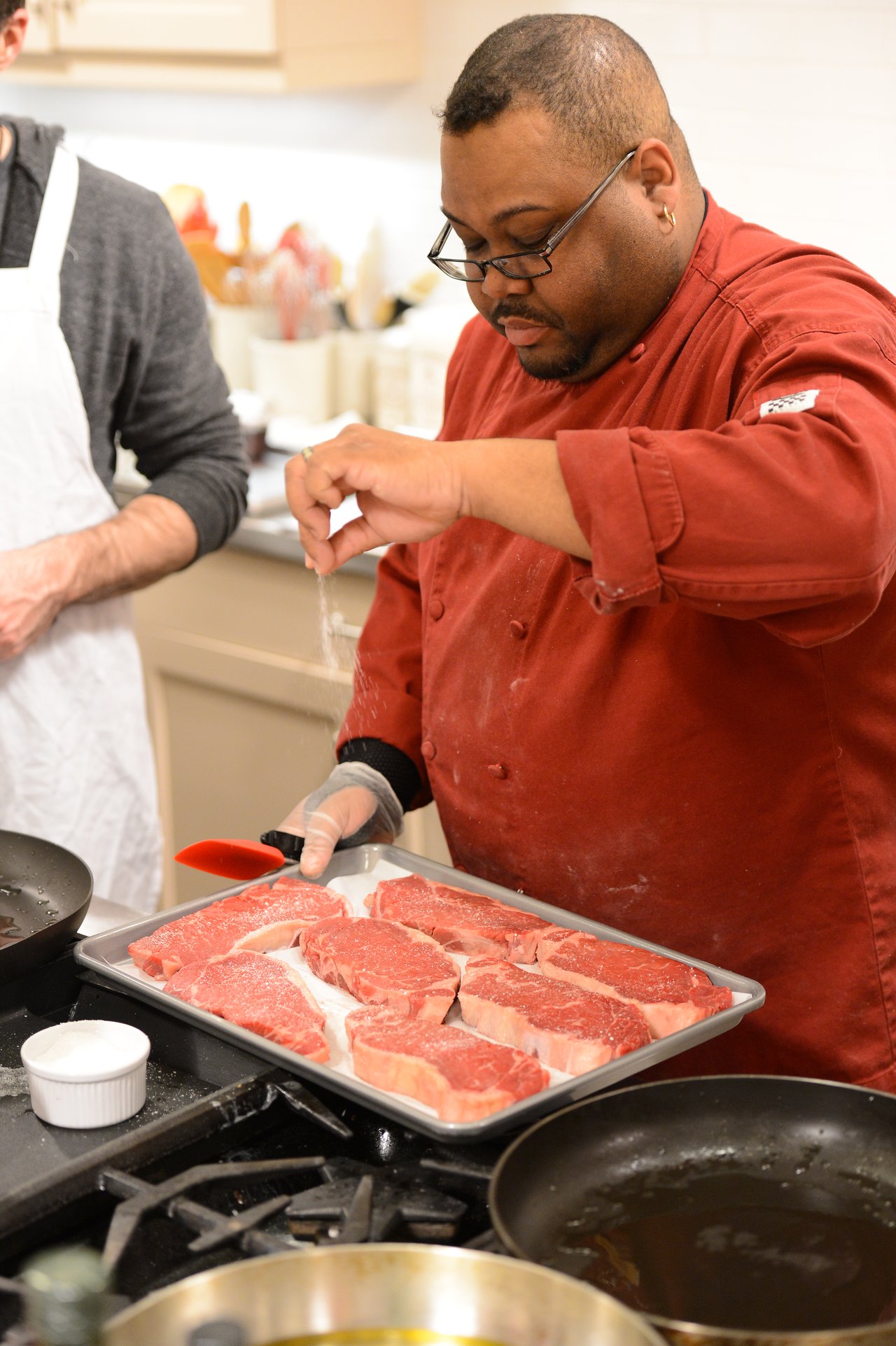 A chef in a red coat sprinkles seasoning on raw steaks placed on a tray in a kitchen.