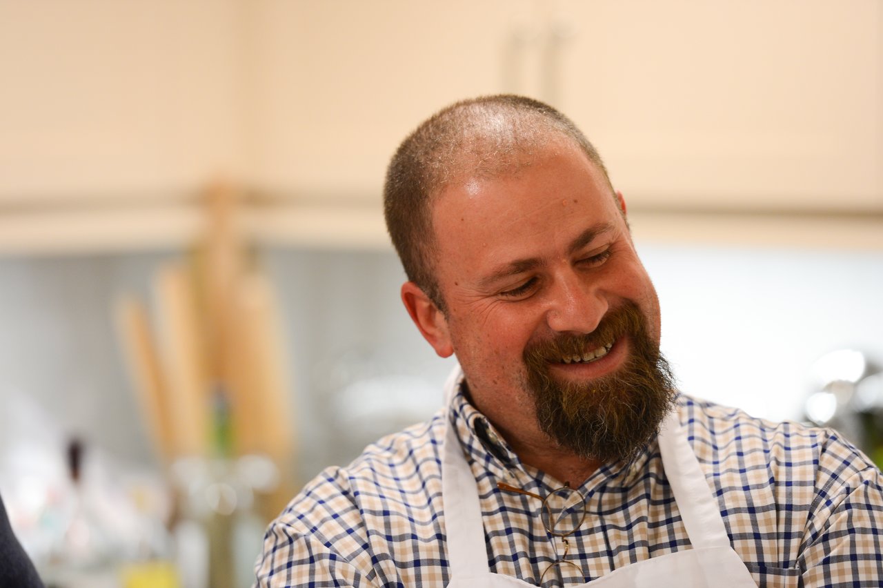 A smiling man wearing a checkered shirt and apron participates in a cooking class in a kitchen setting.