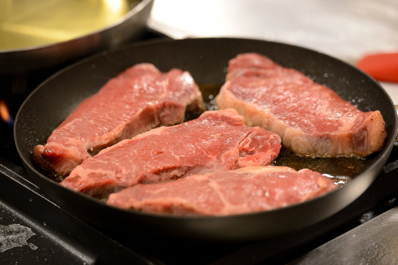 Four raw steaks are sizzling in a black frying pan on a stovetop with oil.