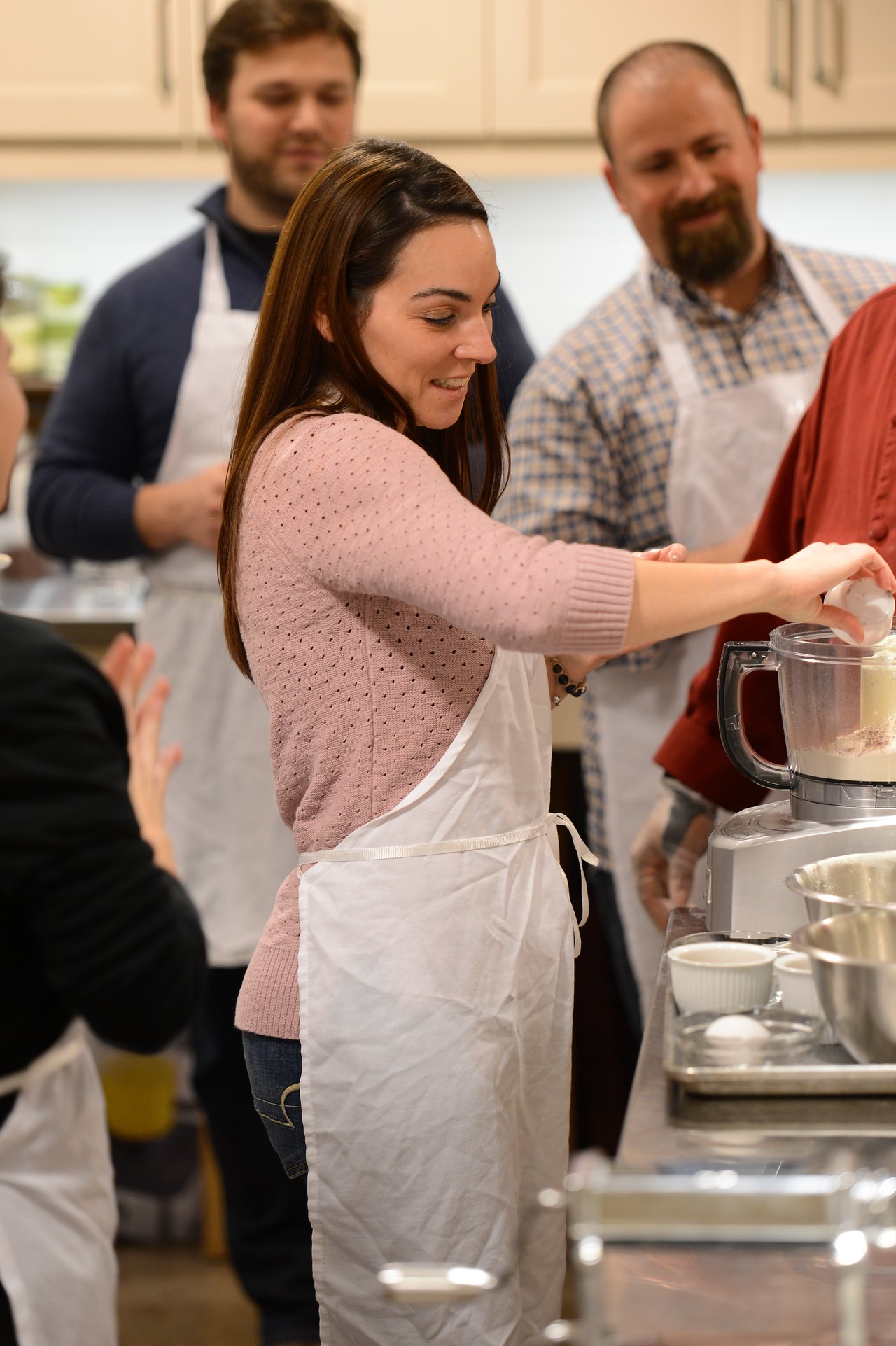 A woman in a white apron pours an ingredient into a food processor while others watch in a cooking class.