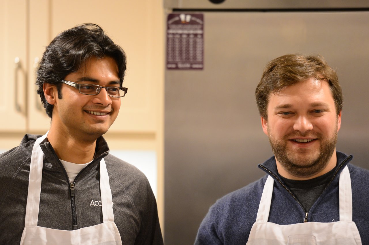 Two people wearing aprons smile in a kitchen, possibly participating in a cooking class.