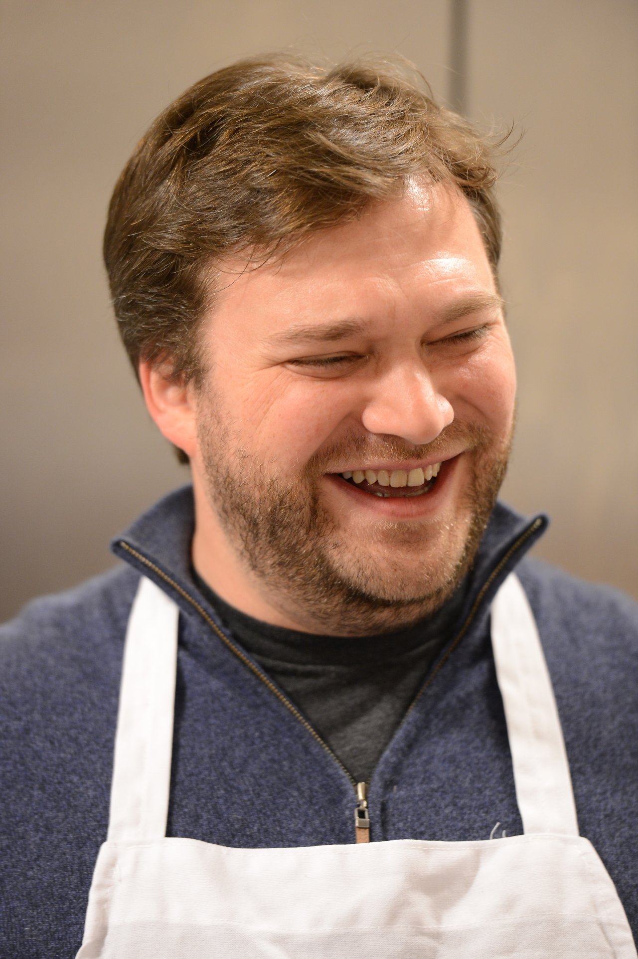A man wearing a white apron smiles and laughs, possibly during a cooking class.