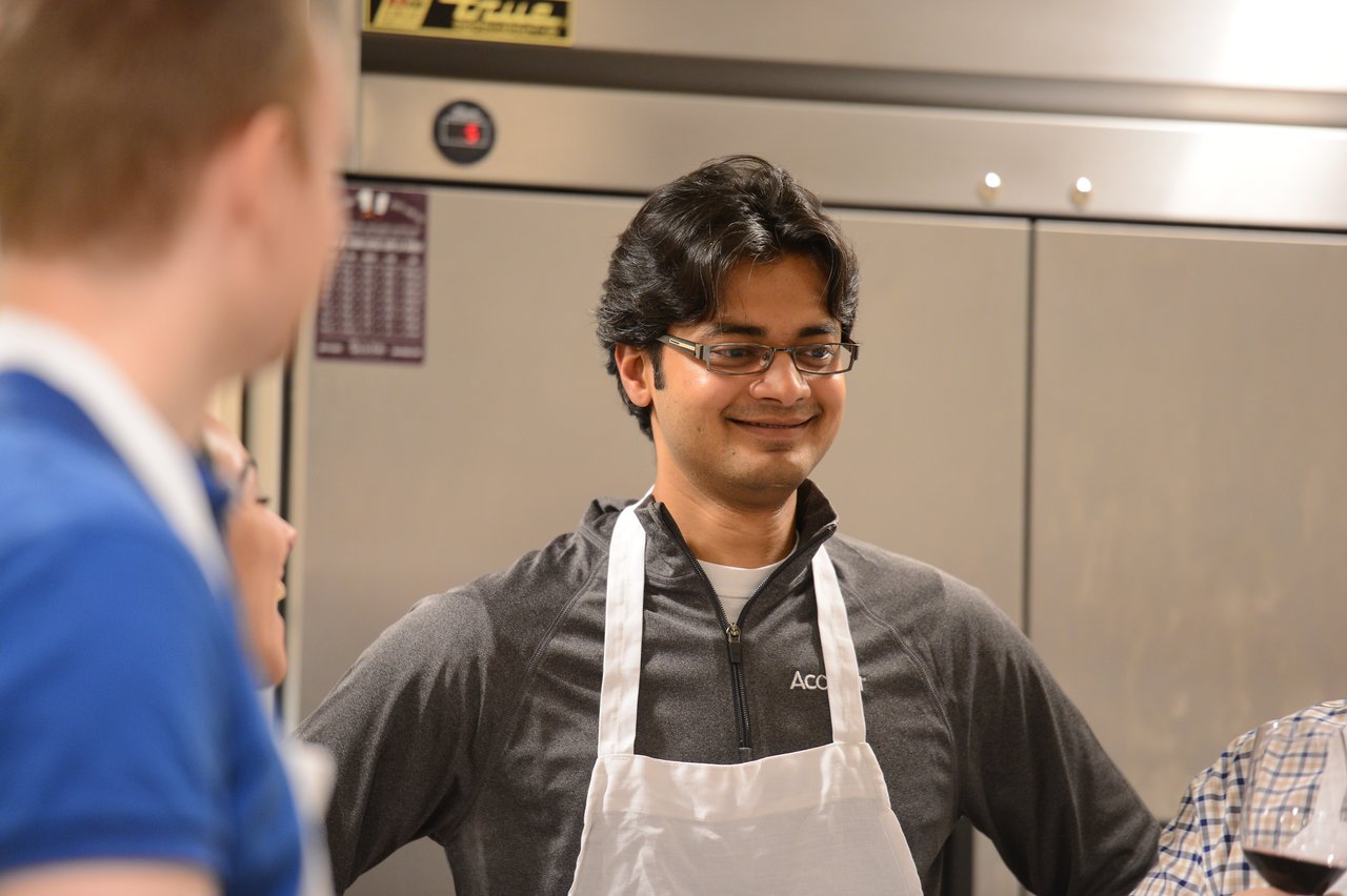 A person wearing an apron smiles while participating in a cooking class, surrounded by others in a kitchen setting.