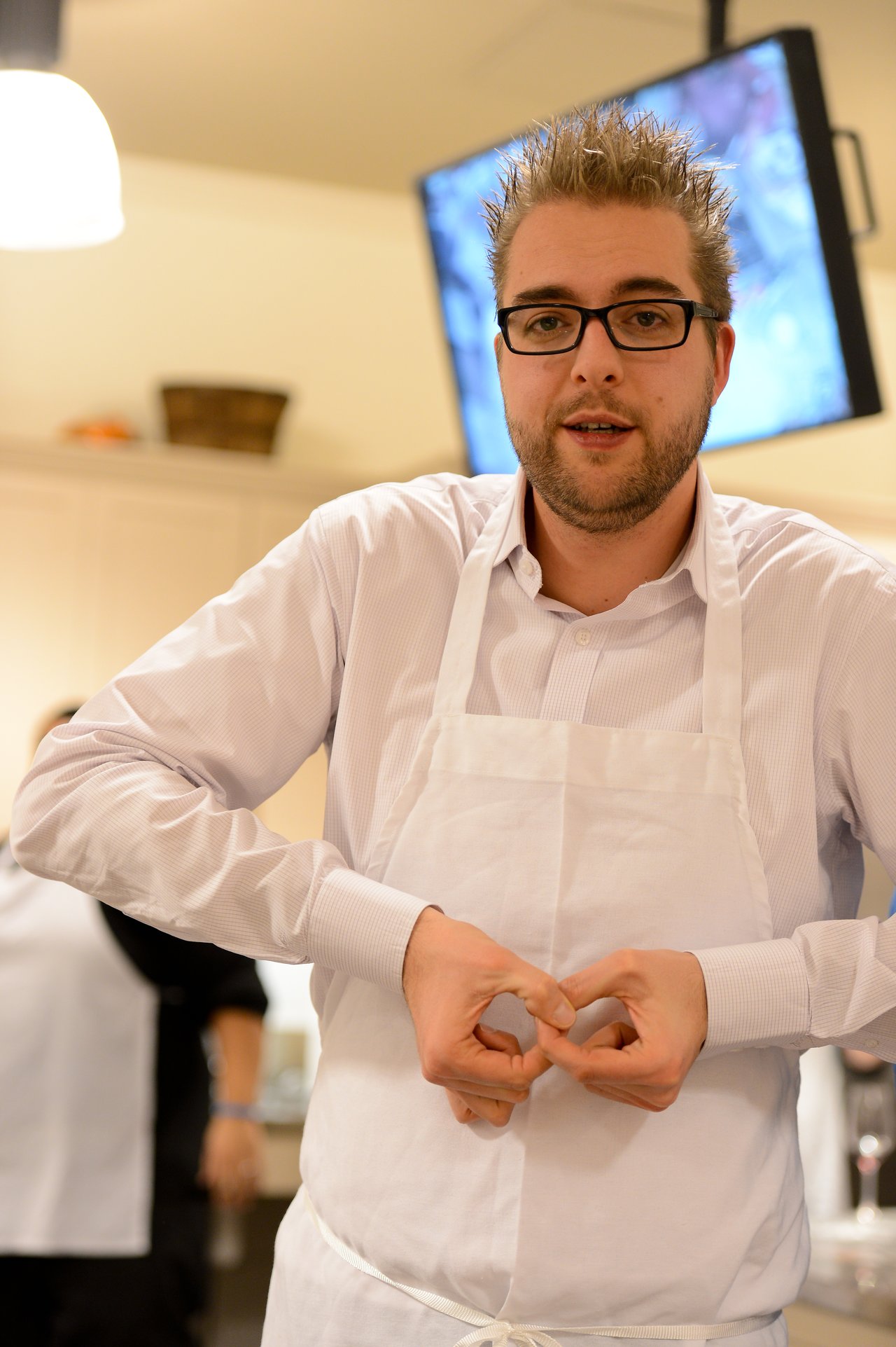 A man wearing glasses and a white apron gestures with his hands during a cooking class.
