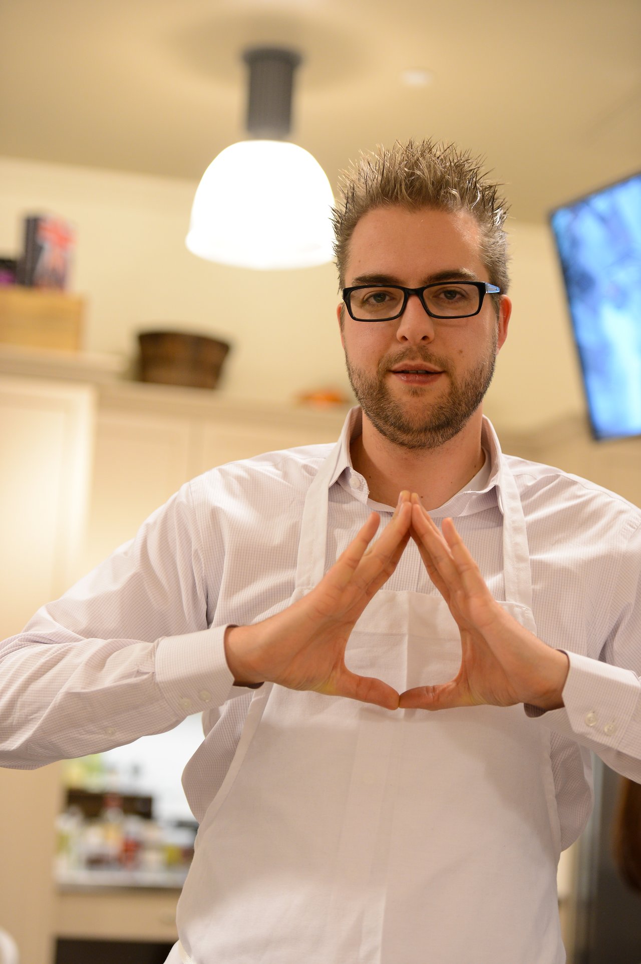 A man wearing glasses and a white apron gestures with his hands in a cooking class setting.