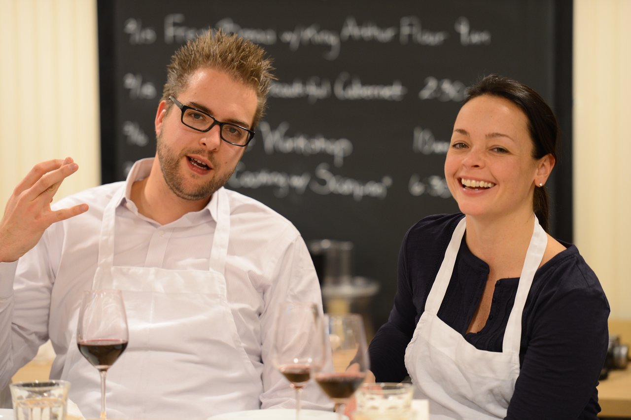 Two people wearing aprons sit at a table with wine glasses, smiling and talking during a cooking class.