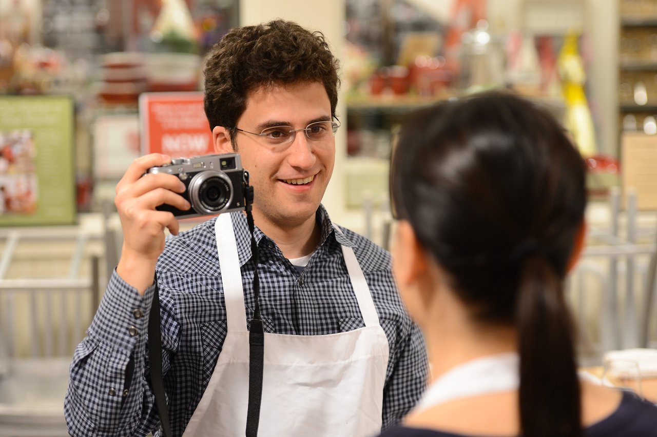 A man wearing an apron holds a camera and smiles while facing a woman in a cooking class.