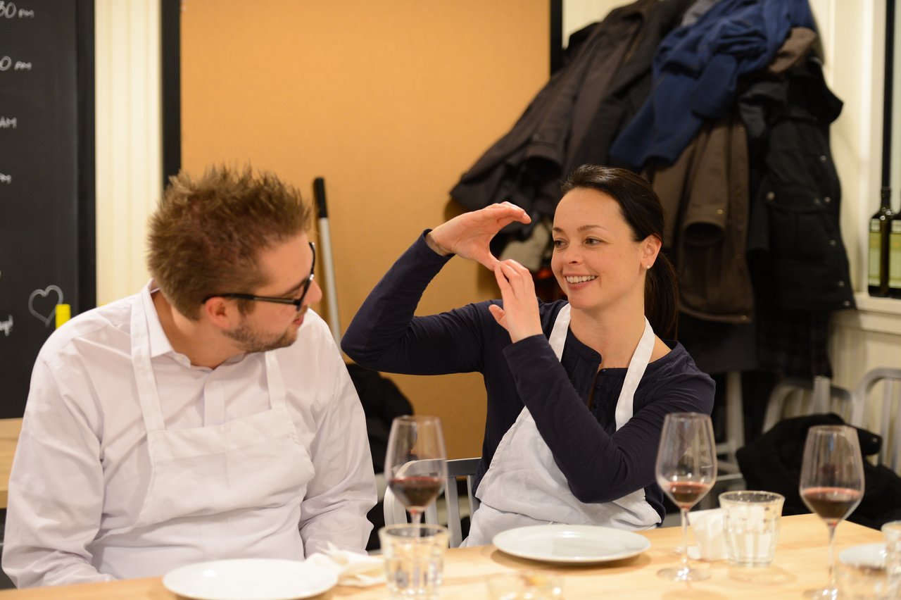 Two people wearing aprons sit at a table with wine glasses, engaged in conversation.