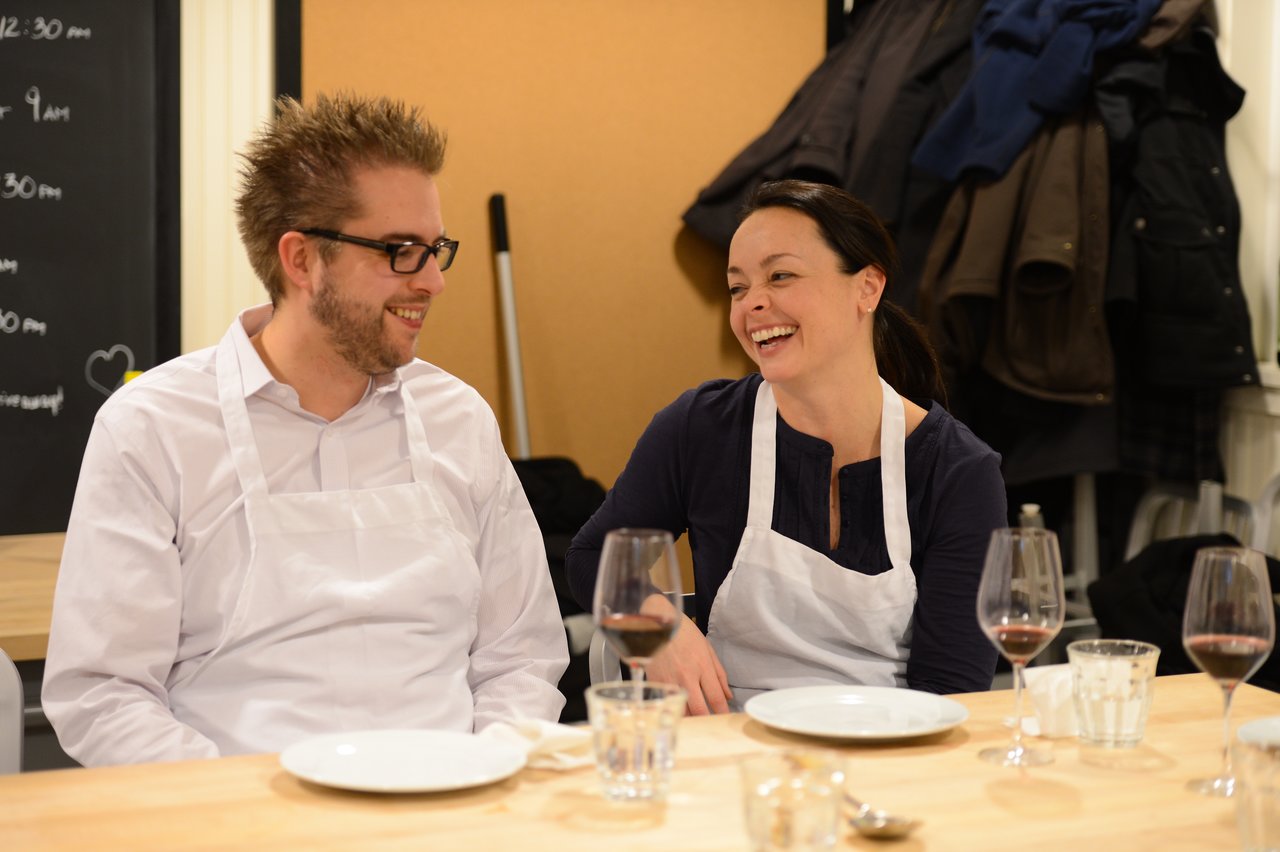 Two people wearing aprons sit at a table, smiling and talking, with plates and wine glasses in front of them.