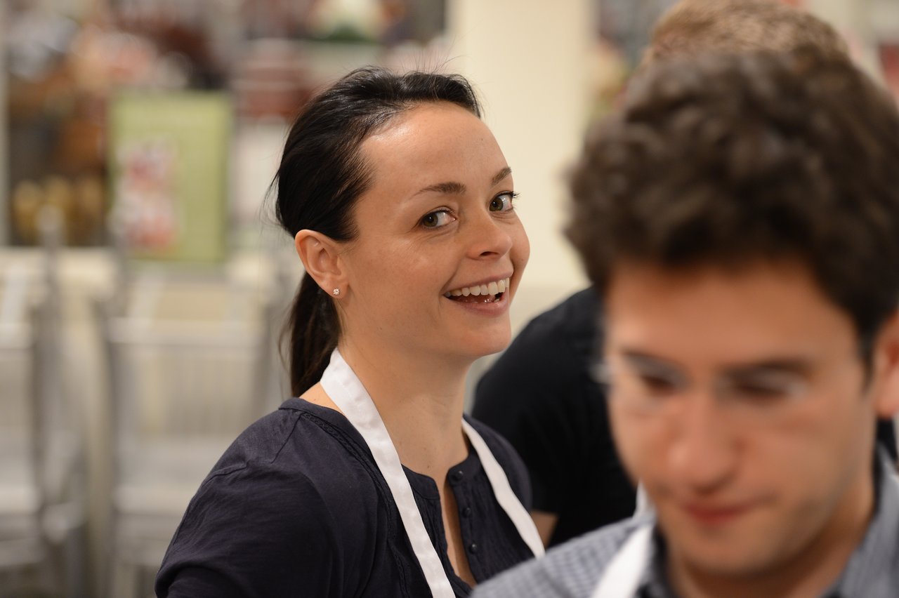 A woman wearing an apron smiles while participating in a cooking class with others in the background.