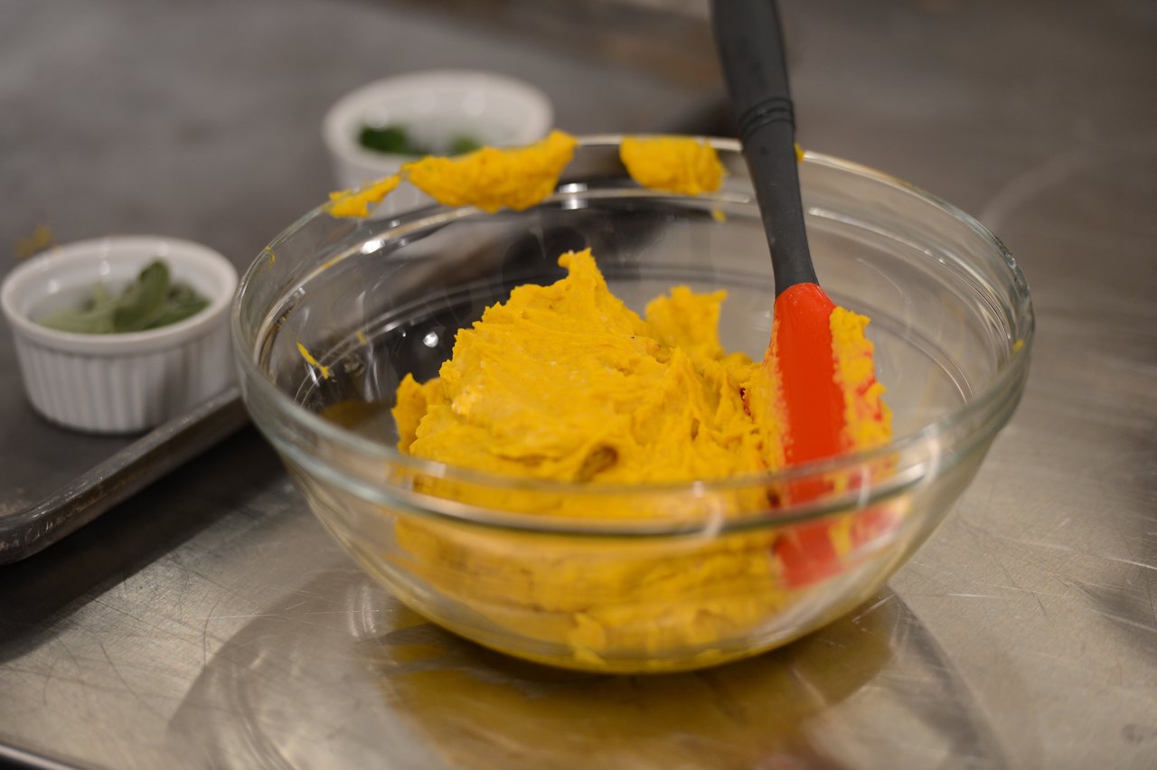 A glass bowl with mashed orange mixture and a red spatula on a stainless steel countertop.