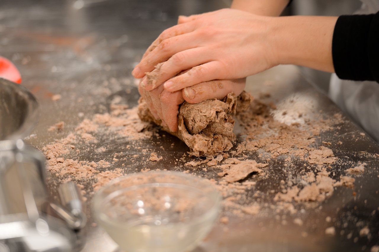 Hands kneading brown dough on a stainless steel surface, with flour and ingredients scattered around.