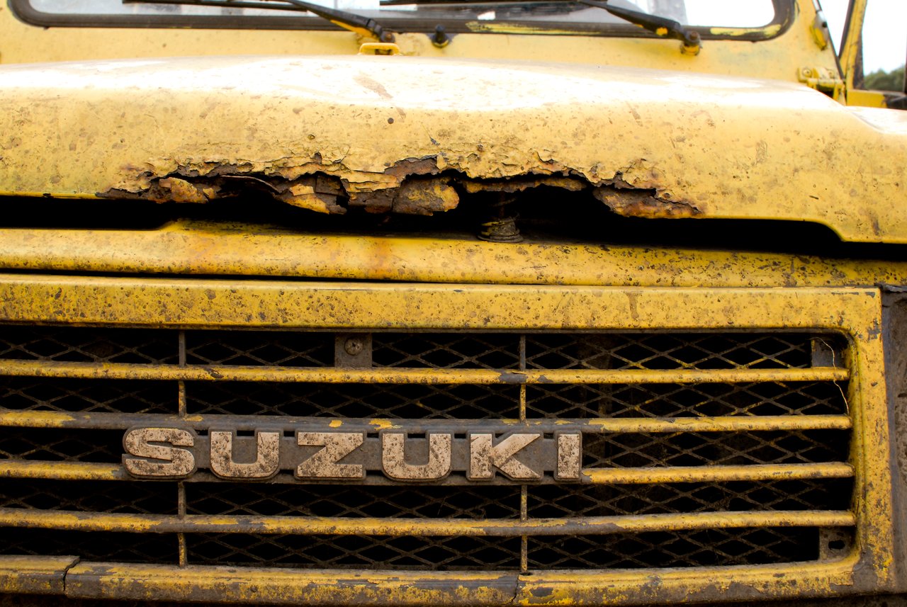 Close-up of a yellow Suzuki vehicle with a rusted and damaged hood, covered in dirt from off-road use.