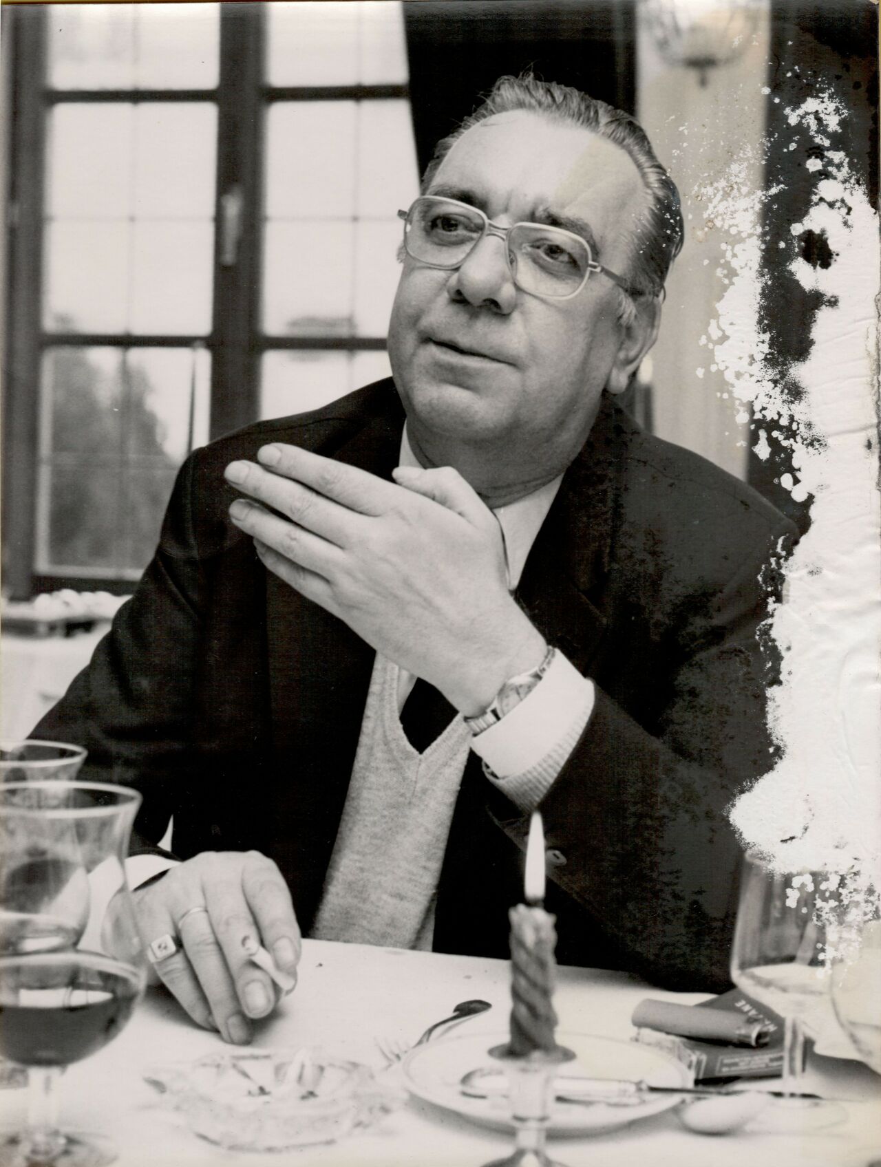 A man wearing glasses and a suit sits at a table, gesturing with one hand while holding a cigarette.