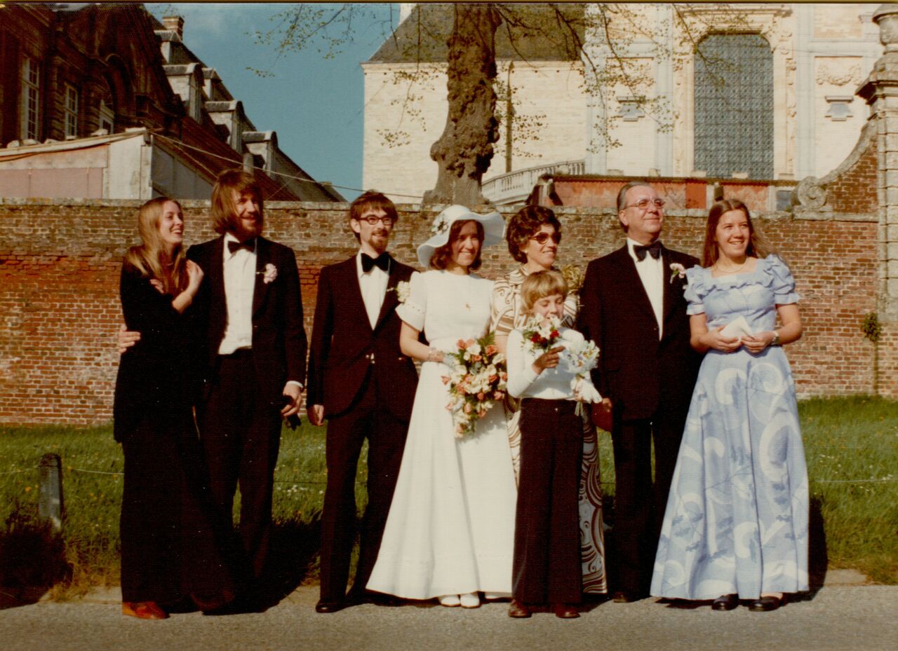 A bride and groom stand with family and friends, dressed in formal attire, posing for a wedding photo outdoors.