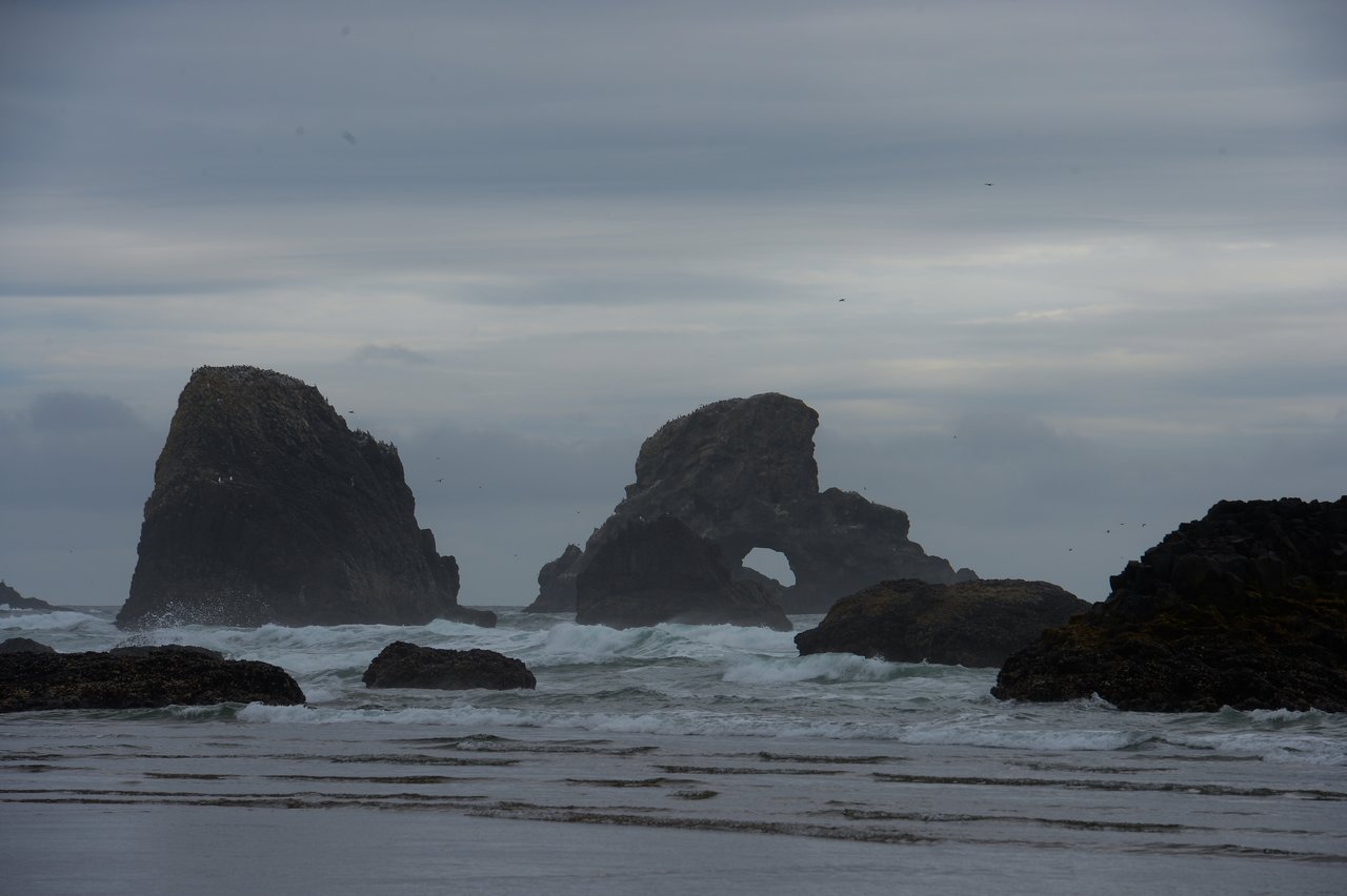 Rock formations rise from the ocean as waves crash against them under a cloudy sky at Cannon Beach.