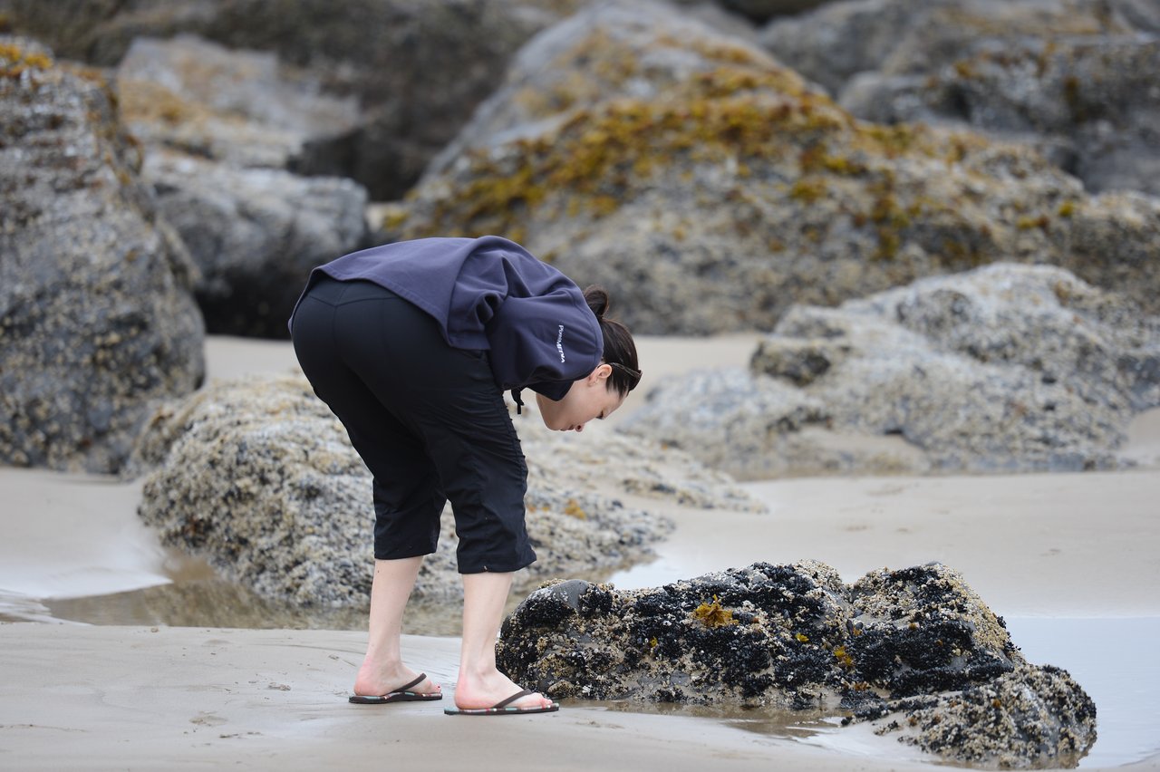 A person in a dark jacket and flip-flops bends down to examine a rock on the beach.