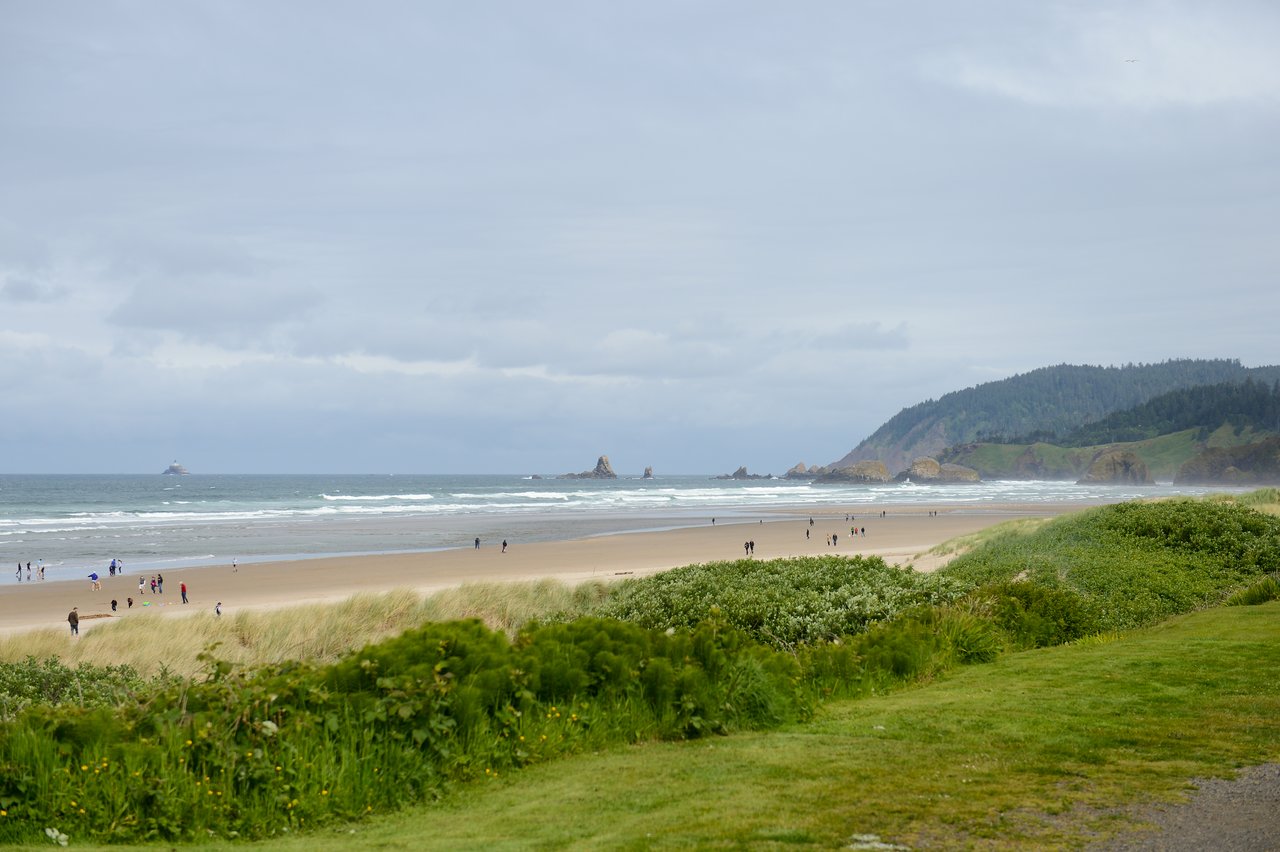 People walking along a wide sandy beach with ocean waves, distant rock formations, and green hills in the background.