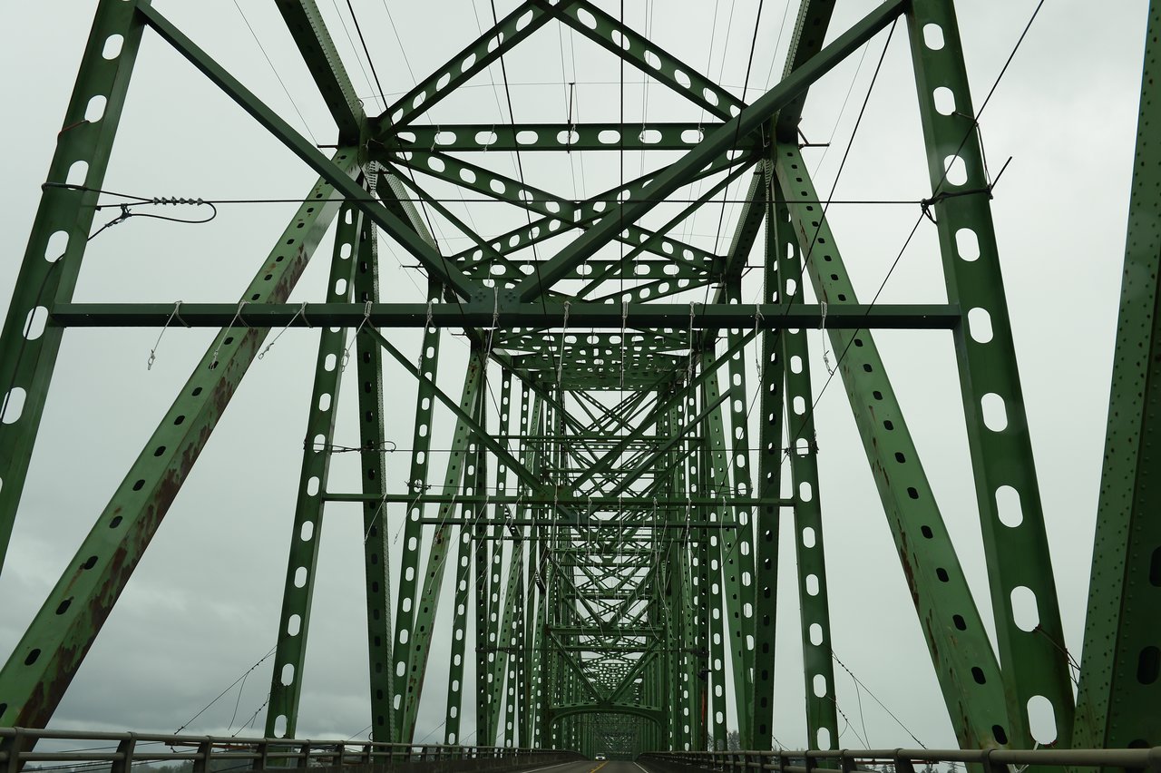 A green metal truss bridge with beams and cables, viewed from the roadway under an overcast sky.