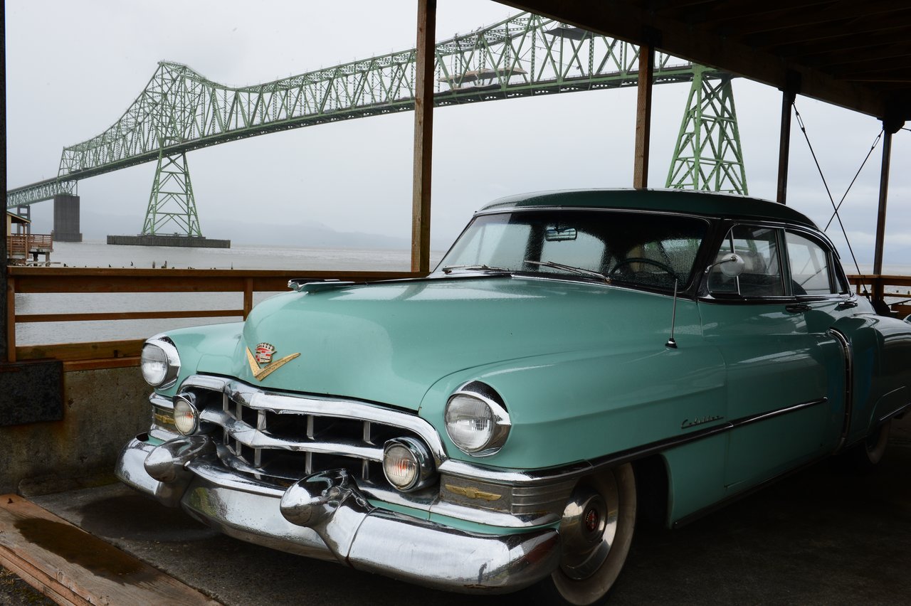 A vintage turquoise Cadillac is parked under a shelter near a large green steel bridge over the water.