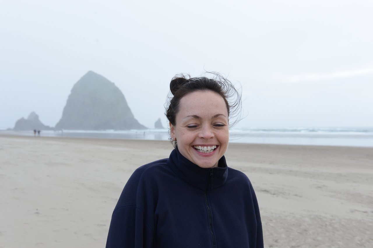 A smiling woman in a dark jacket stands on a sandy beach with large rock formations in the background.