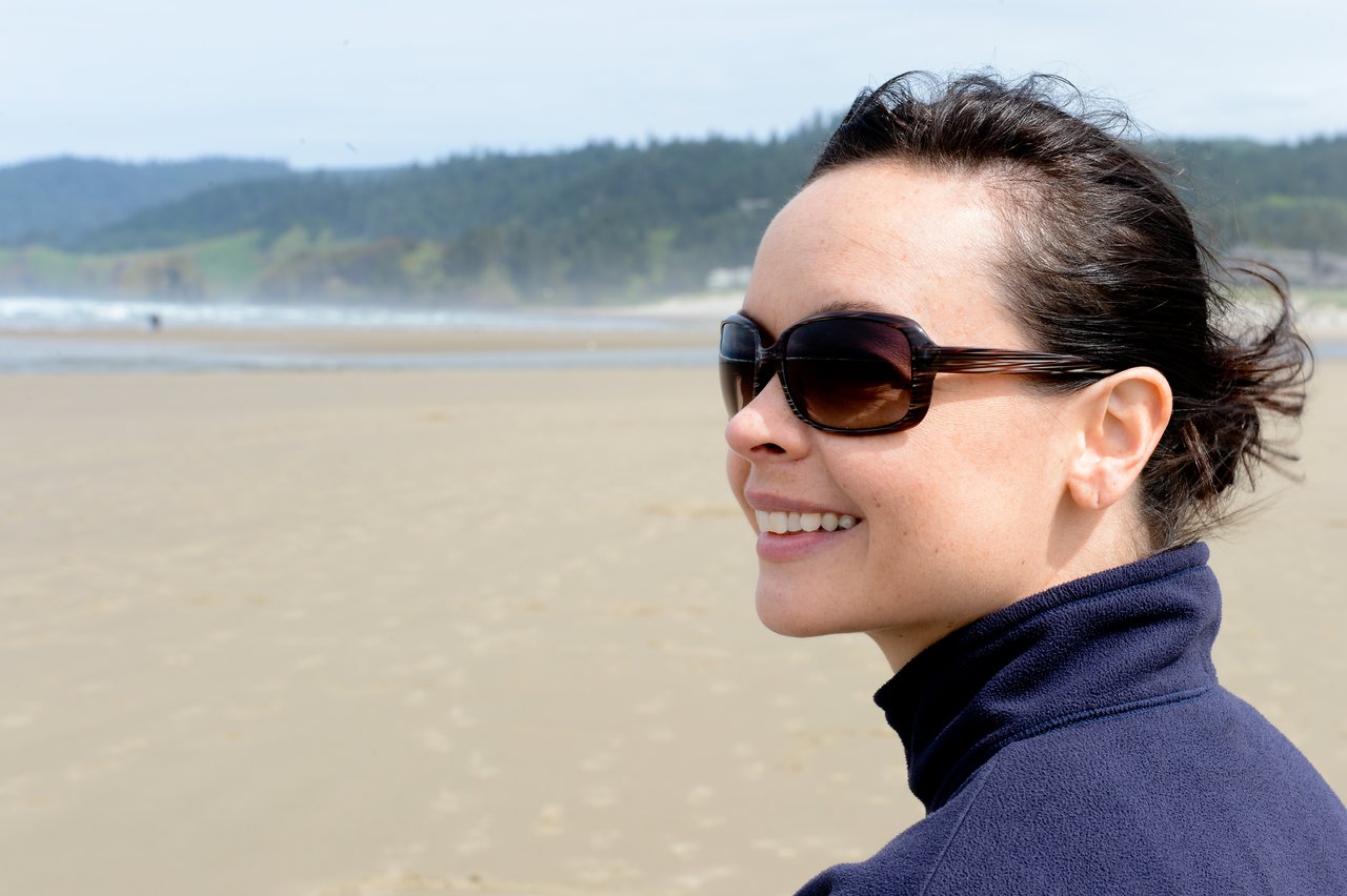 A woman wearing sunglasses and a dark jacket smiles while standing on a sandy beach with ocean waves behind her.