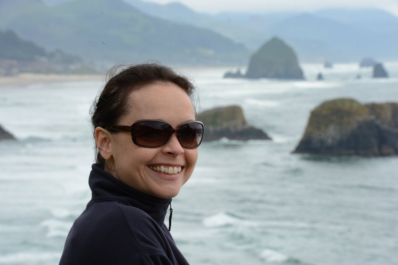 A woman wearing sunglasses and a dark jacket smiles in front of the ocean and rocky formations at Cannon Beach.
