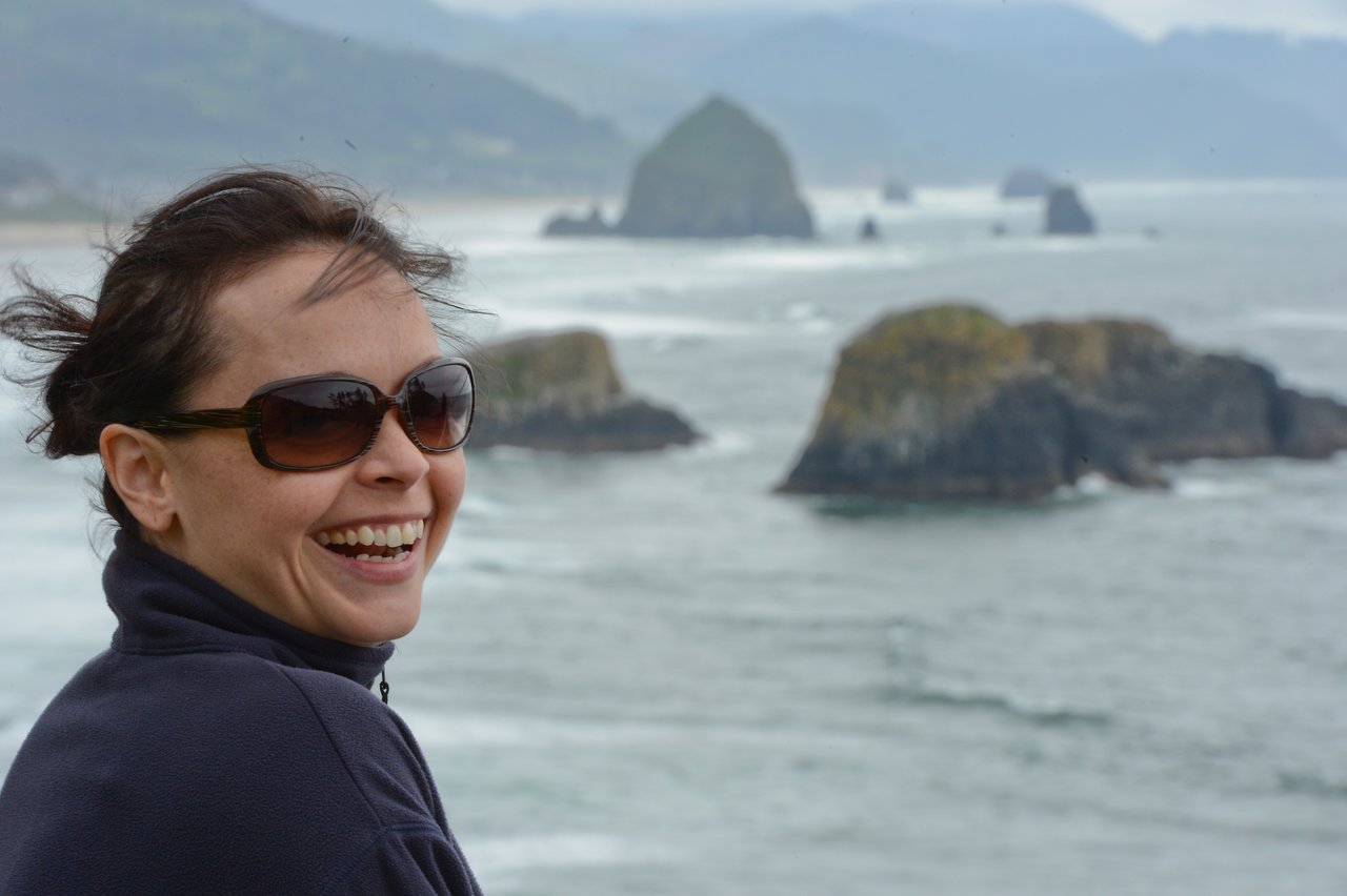 A woman wearing sunglasses and a dark jacket smiles while standing near the ocean with rocky islands in the background.