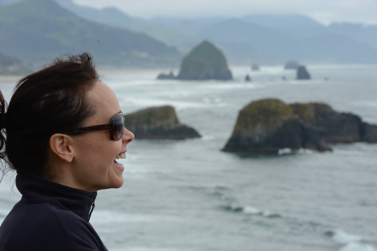 A woman wearing sunglasses and a jacket smiles while looking out at the ocean and rock formations at Cannon Beach.