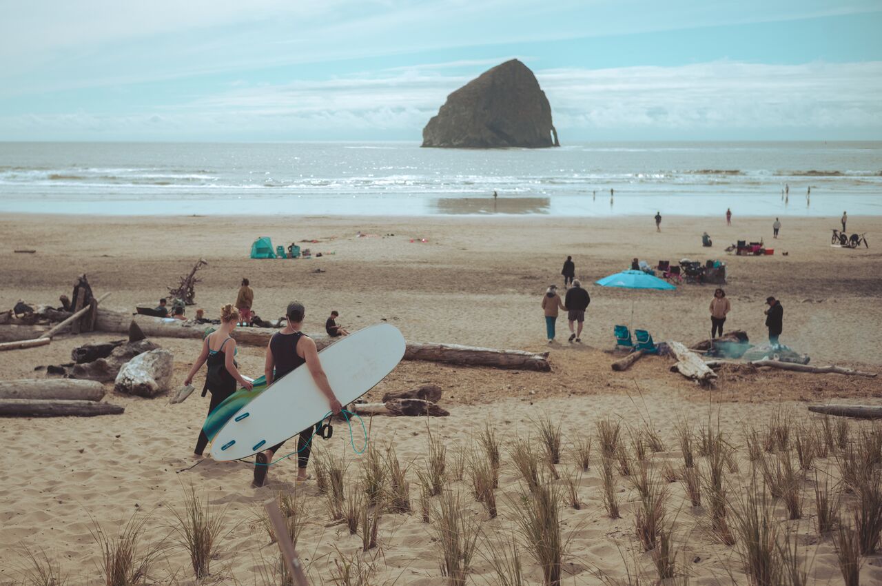 Two people carrying surfboards walk toward the beach with a large rock formation in the water.