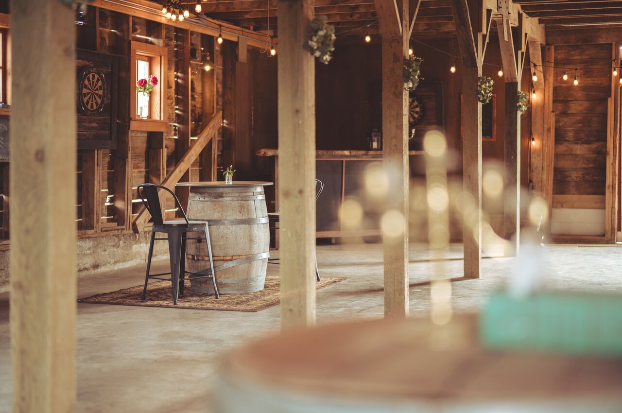 Two chairs sit at a barrel table inside a rustic barn decorated with string lights and flowers.
