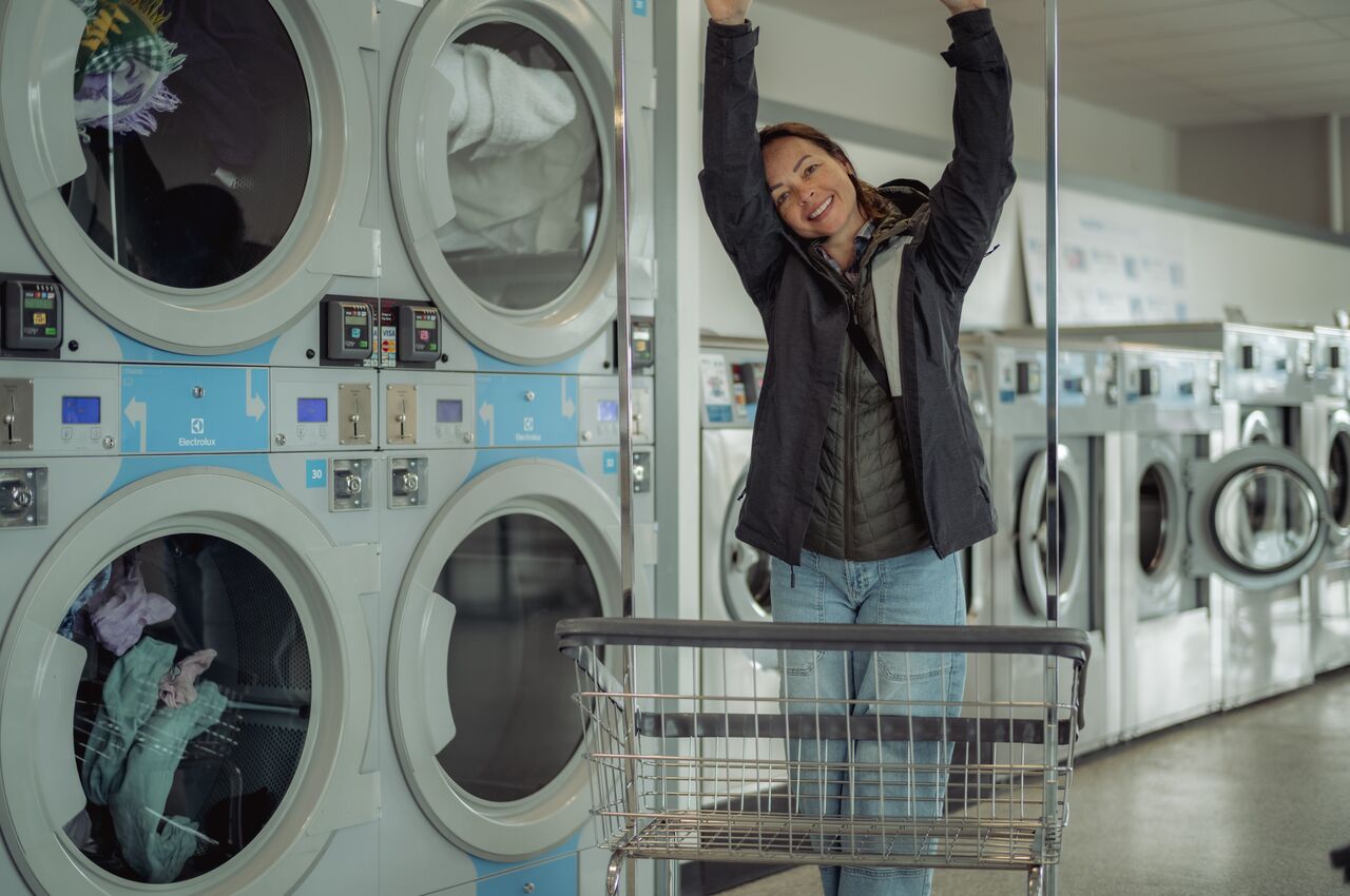 Person stretching and smiling next to a laundry cart in front of dryers at a laundromat.