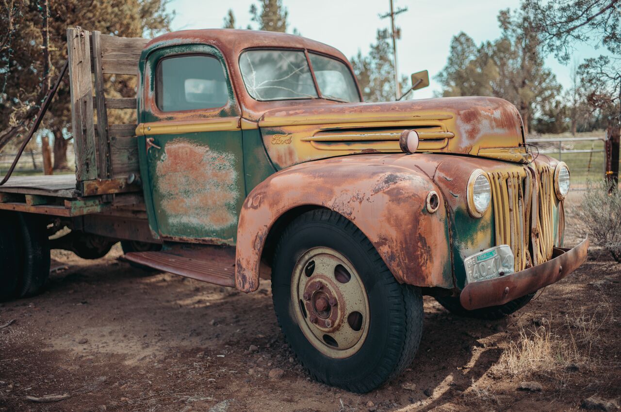 Old rusted Ford truck parked on dirt, with faded paint and wooden flatbed in the back.
