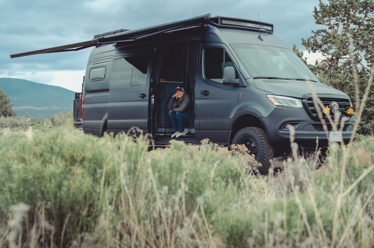 Person sitting in the doorway of a parked camper van at a quiet, remote campground.