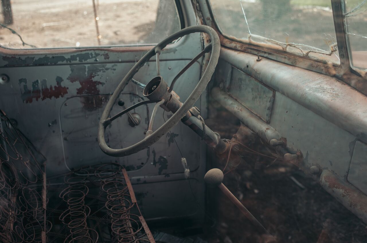 Rusty steering wheel and exposed seat springs inside an old, abandoned truck with cracked windows.