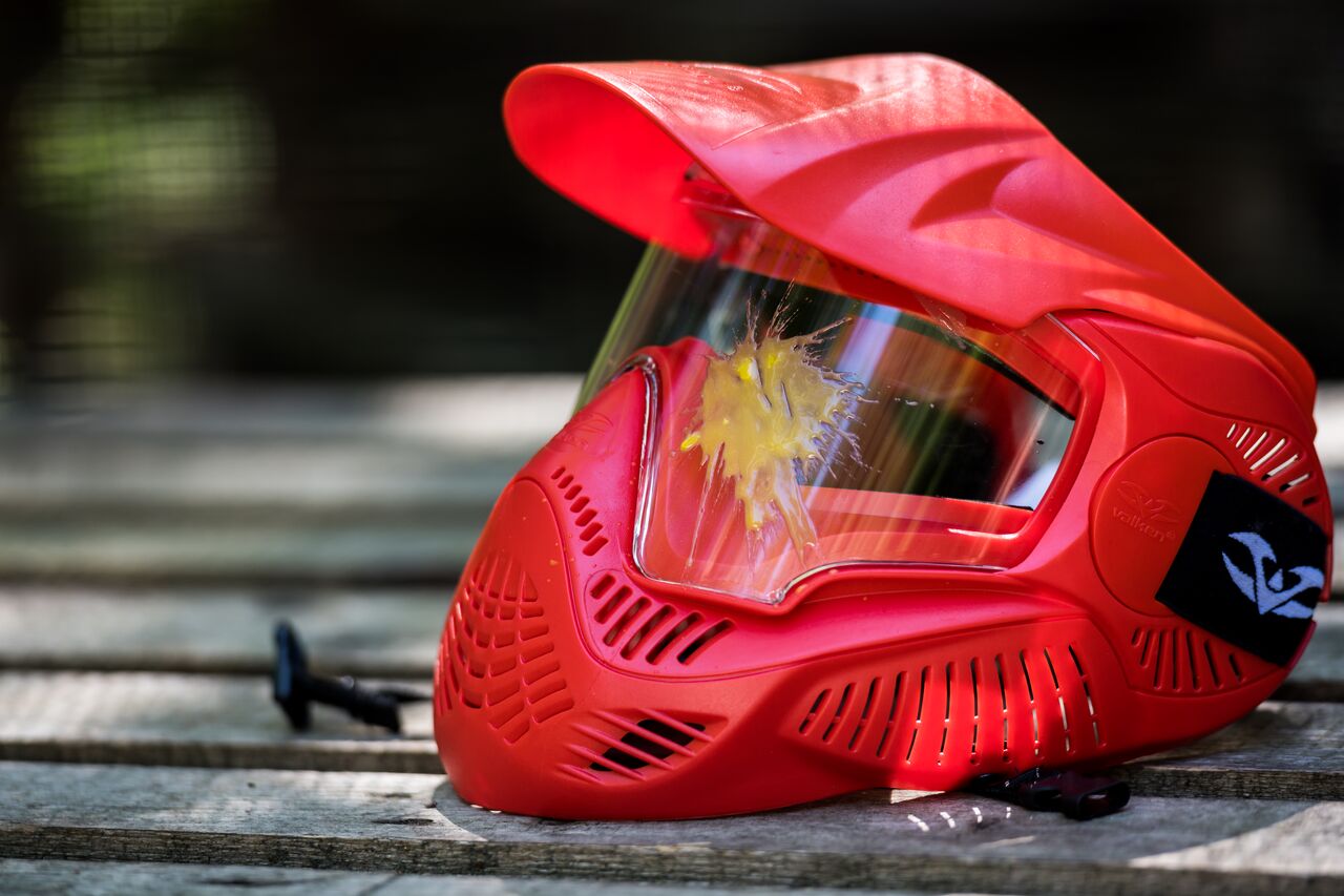 A red paintball mask with a yellow paint splatter on the visor rests on a wooden surface.