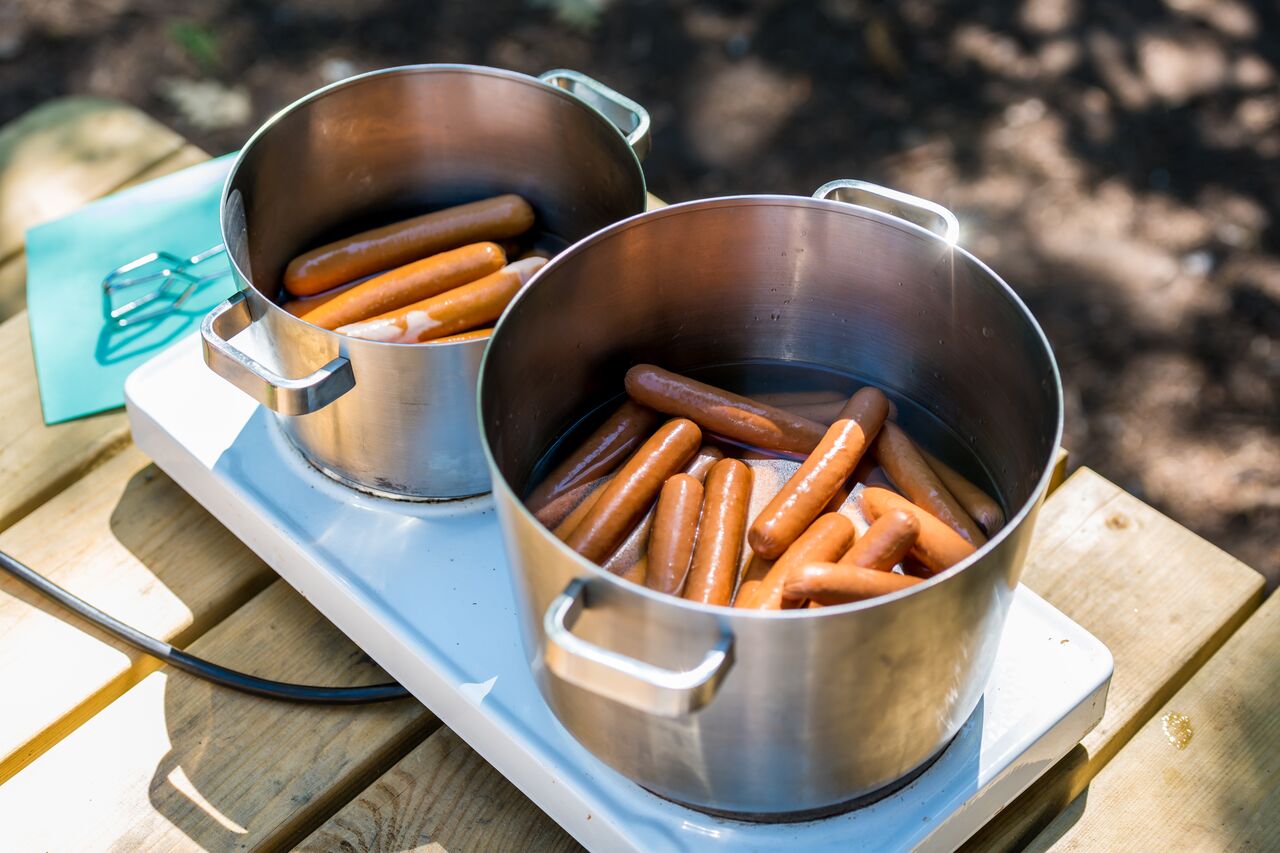 Two large pots of hot dogs boiling on a portable stove outdoors.