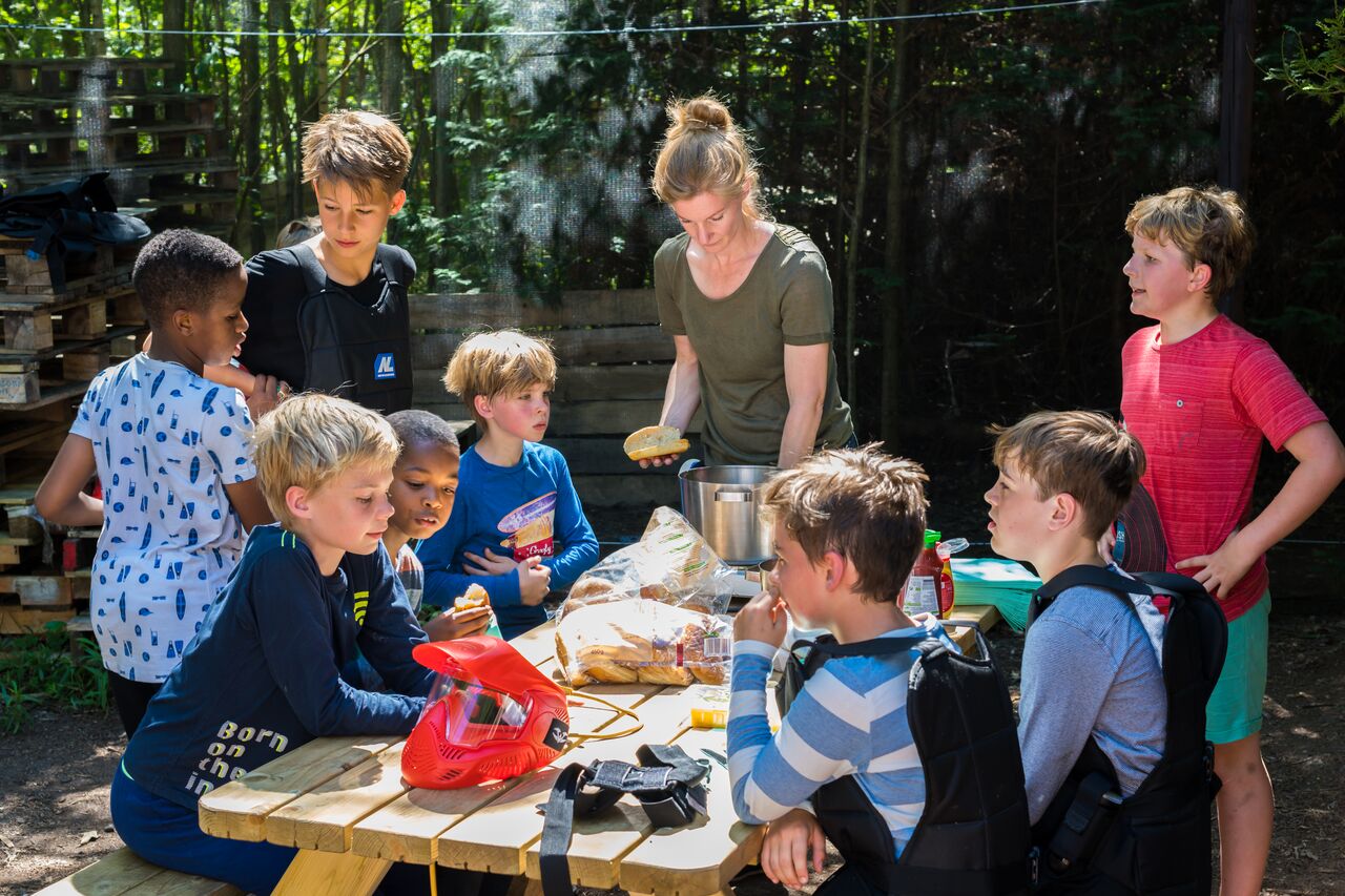 A group of children in paintball gear gather around a table while an adult prepares food outdoors.