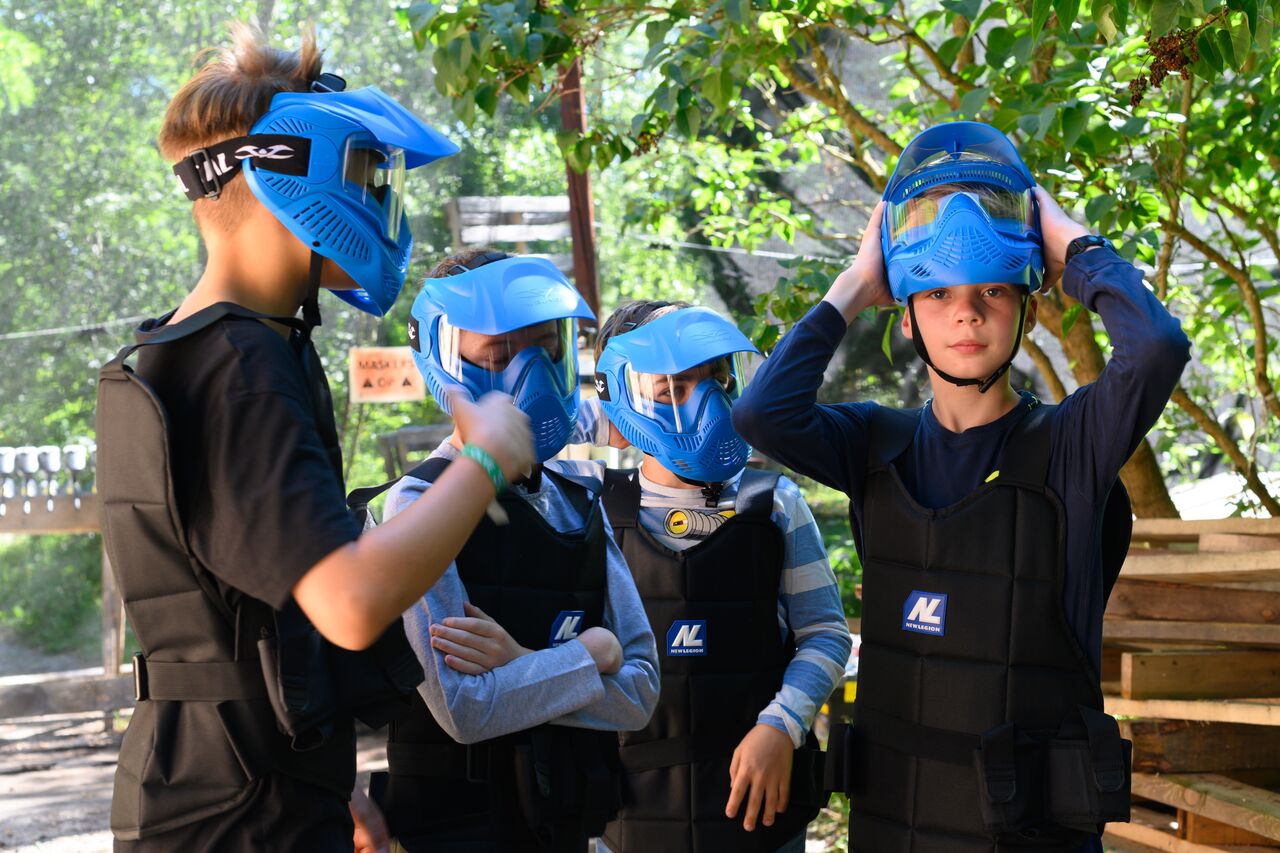 A group of children wearing protective vests and blue paintball masks prepare for a game in an outdoor setting.