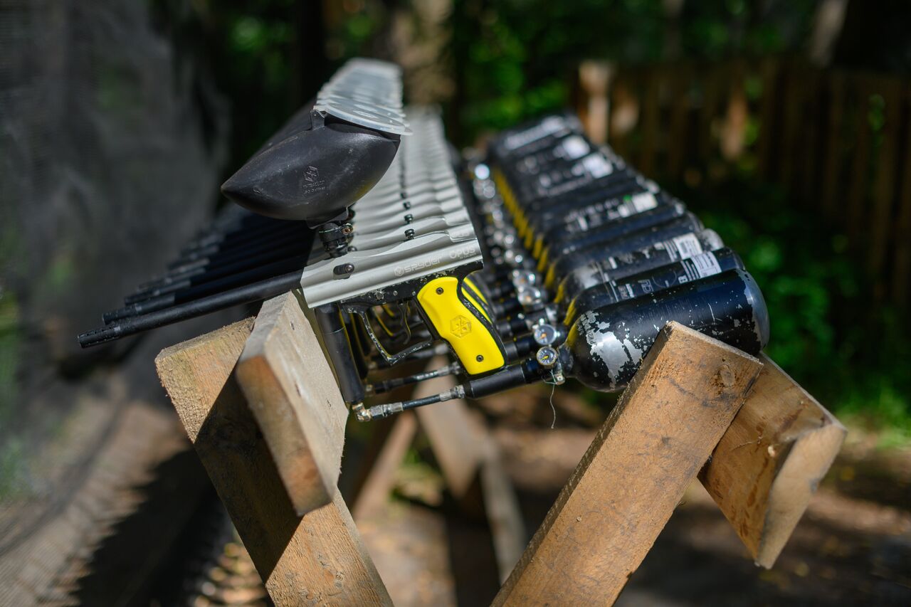 A row of paintball guns with air tanks lined up on a wooden stand outdoors.