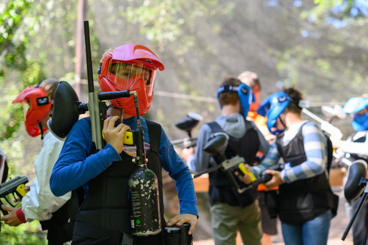 A person in a red helmet holds a paintball gun, surrounded by others preparing for a paintball game.