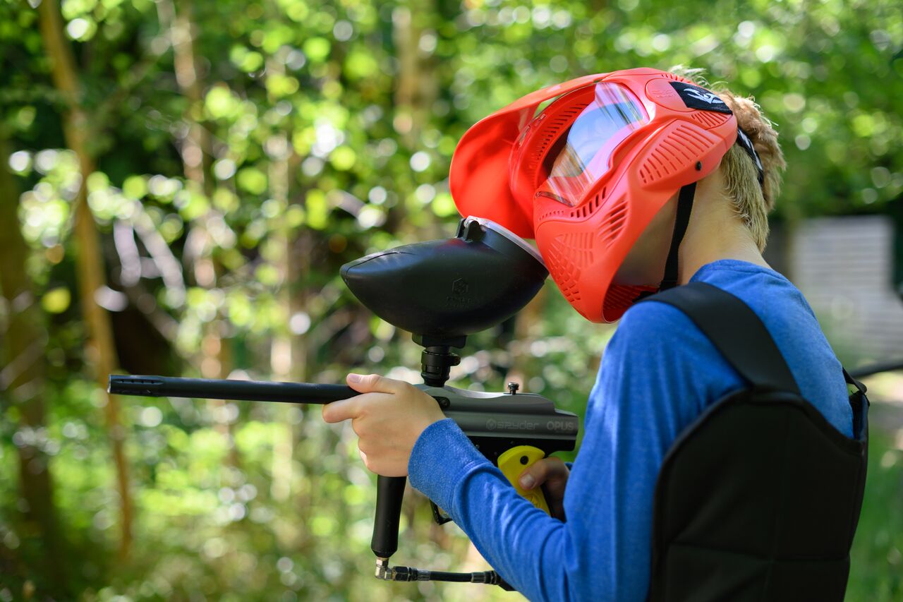 A person wearing a red paintball mask and blue shirt aims a paintball gun in an outdoor setting.