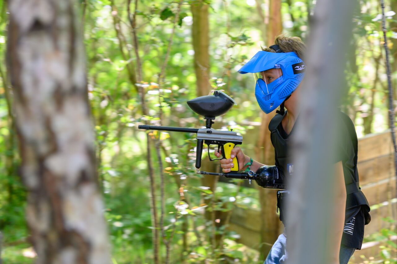 A person wearing a blue paintball mask holds a paintball gun while moving through a wooded area.