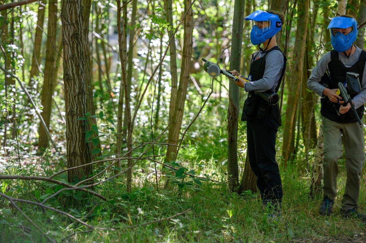 Two people wearing blue helmets and protective vests hold paintball guns while standing in a wooded area.